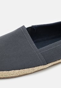Grå lerret slip-on sko med vevet jute såle. Har en teksturert overdel og kontrasterende søm langs kantene.