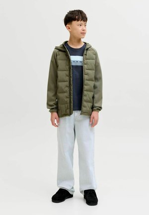 Chaqueta acolchada en color oliva con capucha, diseño acolchado, combinada con una camisa azul oscuro, vaqueros de lavado claro y zapatos negros.