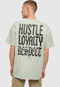 Lysegrøn oversized t-shirt med fed sort typografi på ryggen: "HUSTLE LOYALTY RESPECT", med markante, stiliserede skrifttyper.