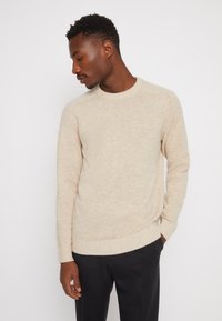 Beige strikkegenser med rund hals, ribbestrikkede ermer og kant. Myk tekstur, normal passform, brukt sammen med mørke bukser.