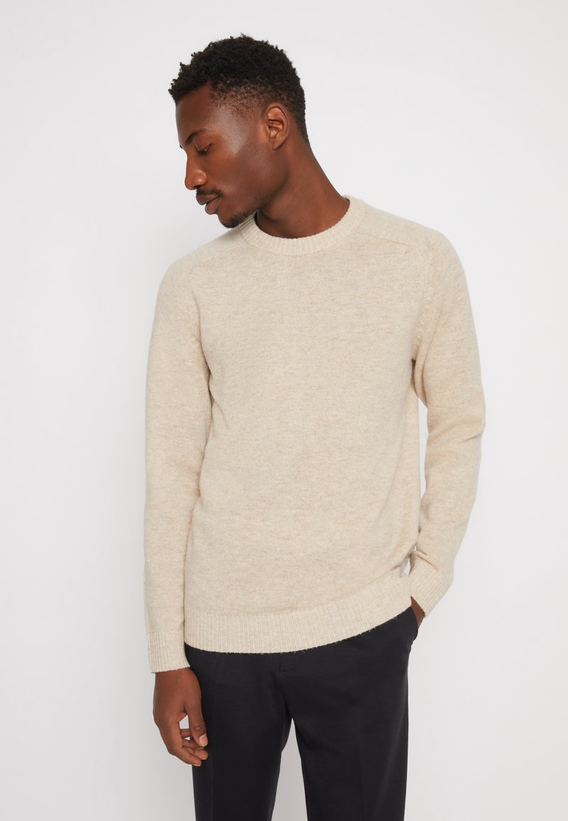 Beige strikkegenser med rund hals, ribbestrikkede ermer og kant. Myk tekstur, normal passform, brukt sammen med mørke bukser.
