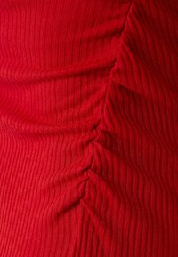 Röd ribbad tyg med vertikala linjer och en rynkad detalj på ena sidan, som visar en texturerad yta och en subtil lyster.