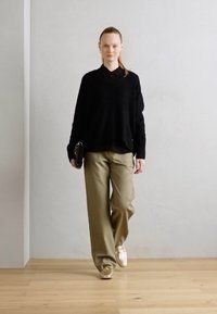 Pull noir surdimensionné, chemise sombre en dessous, pantalon large beige, et baskets dorées. Une mannequin tenant une pochette noire marche sur un sol en bois.