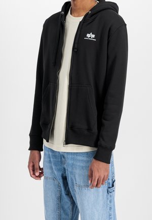 Mężczyzna w czarnej bluzie Alpha Industries zapinanej na zamek, jasnobeżowej koszuli oraz jasnoniebieskich jeansach z kieszeniami, stojący na białym tle.