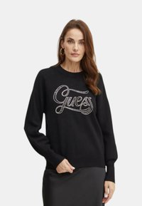 Maglione nero con logo "Guess" in filo metallico. Maniche lunghe con polsini aderenti. Tessuto morbido e design morbido.
