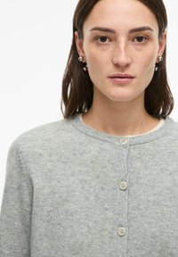 Mlada ženska z rjavimi očmi in pegicami, ki nosi srebrne uhane in svetlo sivi gumbasti kardigan čez belo majico na belem ozadju.