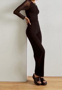 Jumpsuit noir ajusté avec des manches longues en tulle, fermeture éclair et col plat. Assorti avec des sandales à plateforme chunky. Texture lisse, accents minimaux.