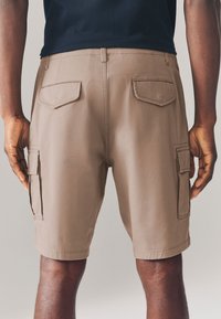 Shorts cargo marron avec poches à l'avant et à l'arrière, présentant un design épuré et une texture lisse. Le tissu semble léger et durable.