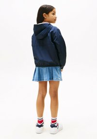 Veste à capuche bleu marine avec des poignets élastiques, portée sur une jupe en denim clair. Le modèle l'associe à des baskets blanches et des chaussettes à rayures rouges.