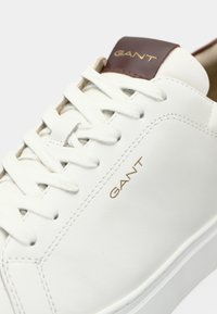 Sneaker alb din piele, cu sireturi albe plate, având un accent din piele maro pe toc și un branding aurit "GANT" pe lateral.