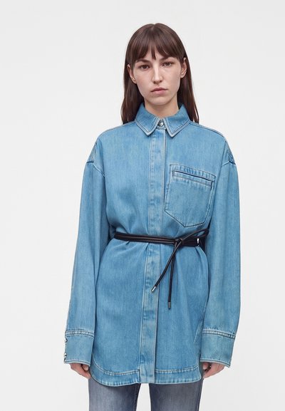 Jeune femme aux cheveux bruns et lisses portant une chemise en denim bleu clair surdimensionnée, cintrée à la taille avec une fine ceinture noire.