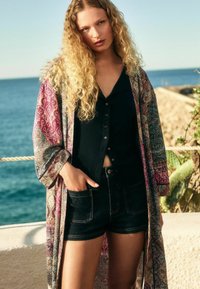 Rakstains garš kimono spilgtā rozā, violetā un zilā krāsā; kombinēts ar melnu pogu augšu un tumšiem džinsu šortiem. Redzami teksturēti auduma detalizējumi.