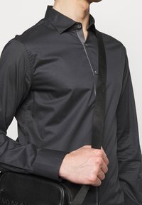 Svart skjorta med knappar och grå detaljer, med långa ärmar och en strukturerad krage. Modellen håller en svart väska med axelrem.