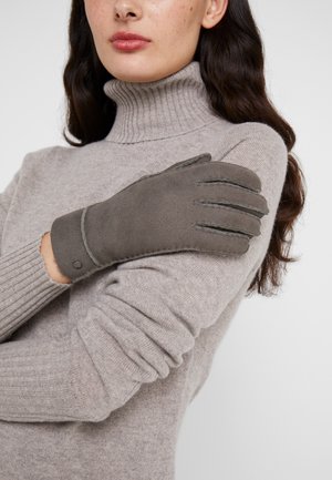 Graue Lederhandschuhe mit fünf Fingern, ausgestattet mit einem gestickten Design und einem Druckknopfverschluss, getragen über einem hellgrauen Rollkragenpullover.