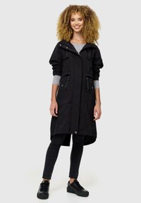 Abrigo negro impermeable con capucha, que cuenta con bolsillos con cremallera, lazos ajustables en la cintura y un corte hasta la rodilla. Lleva puesto sobre una blusa gris y pantalones negros.