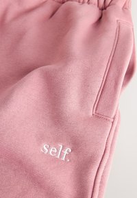 Pantalon de survêtement en coton rose avec une taille élastique, une poche latérale et un logo "self." brodé en blanc près de l'ourlet. Texture lisse.