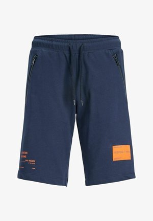 Junge trägt ein blaues T-Shirt mit Brusttasche, dunkelblaue Shorts mit orangefarbenen Aufnähern, weiße Socken und weiße Sneaker und steht mit den Händen in den Taschen.