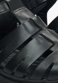 Czarne skórzane sandały z plecionymi paskami, o teksturowanej powierzchni i antypoślizgowej podeszwie z zygzakowatym wzorem na krawędzi.