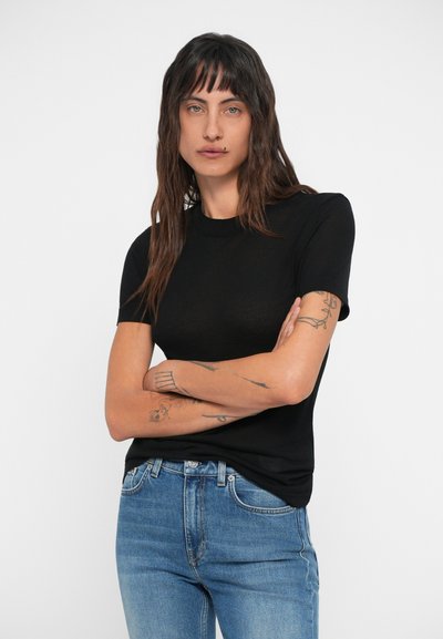 Tricou negru cu mânecă scurtă și decolteu rotund, fabricat dintr-un material moale. Modelul îl poartă cu blugi din denim albastri, eviden�țiindu-se tatuaje subtile pe brațe.
