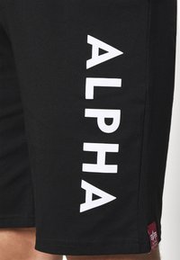 Svart shorts med en slät textur har stora vita "ALPHA"-bokstäver på sidan och en liten vinröd logotyp vid nederkanten.