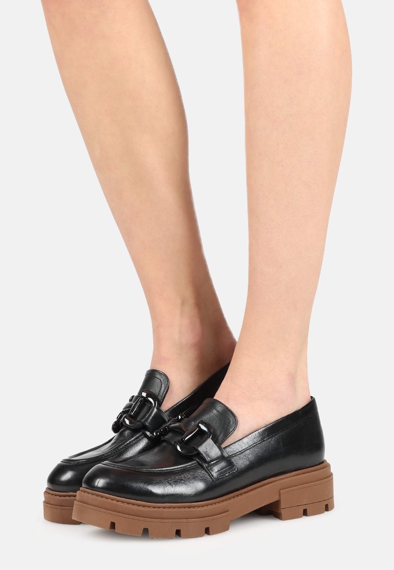 Mocassins en cuir noir avec semelles crantées marron épaisses portés pieds nus sur un fond clair uni.