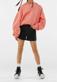 Sudadera oversized de color melocotón con un bolsillo frontal, combinada con pantalones cortos de mezclilla negros. Zapatillas altas negras y calcetines blancos de caña corta visibles.