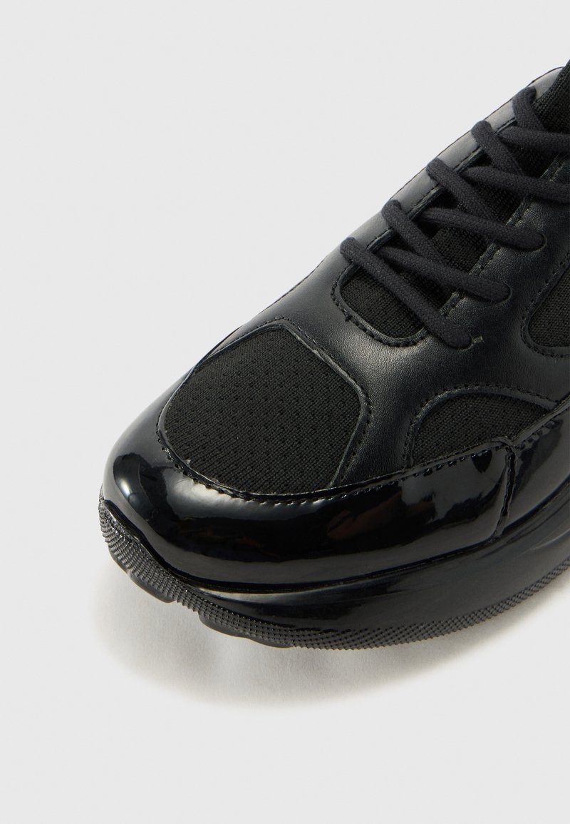 Sneaker nera caratterizzata da una combinazione di pelle verniciata lucida e tessuto in rete testurizzato. Punta rotonda, design con lacci e suola in gomma a contrasto.