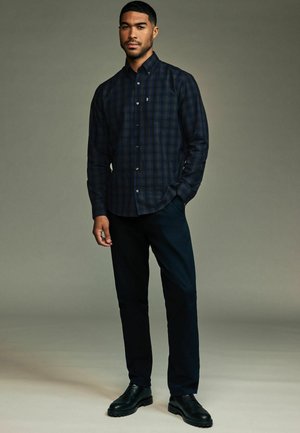 Navy- und grünkariertes Button-Down-Hemd mit langen Ärmeln, kombiniert mit dunklen Hosen und schwarzen Schnürschuhen, vor einem grauen Hintergrund.