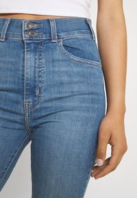 Primer plano de una persona que lleva vaqueros de denim azul de talle alto con doble botón y una camiseta blanca, con la mano relajada a su lado.