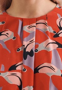 Röd tyg med ett djärvt mönster av abstrakta flamencok inspiration fåglar i svart, rosa och lavendel. Plisserad halsringning ger textur.