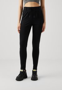 Sorte, tettsittende joggebukser med elastisk midje og snøring; kombinert med store sorte sneakers med gule detaljer.