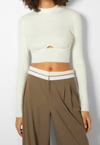 Bílý žebrovaný cropped top s dlouhým rukávem a designem vlny v oblasti pasu, doplněný hnědými plisovanými kalhotami s vysokým pasem.