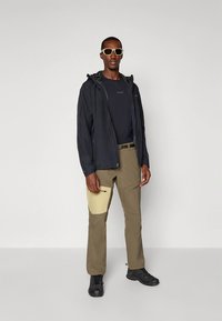 Vandtæt sort jakke, marineblå t-shirt, khakifarvede cargobukser med gul accentlomme, sort fodtøj og hvide solbriller.