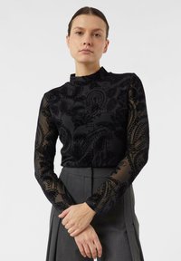 Kobieta w ciemnej przezroczystej bluzce z długim rękawem i wysokim kołnierzem, ubrana w szarą plisowaną spódnicę, stojąca z założonymi rękami na tle białego tła.