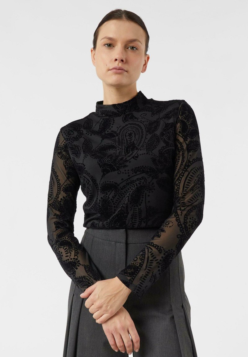 Kobieta w ciemnej przezroczystej bluzce z długim rękawem i wysokim kołnierzem, ubrana w szarą plisowaną spódnicę, stojąca z założonymi rękami na tle białego tła.