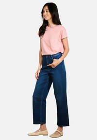 T-shirt rosa a maniche corte abbinato a jeans a vita alta blu scuro e scarpe slip-on beige, su uno sfondo bianco semplice.