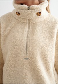 Krémový fleecový pulovr s vysokým límcem, polo-zip uzávěrem a dvěma dekorativními knoflíky. Měkká textura a volný střih. Stříbrné detaily kovu.