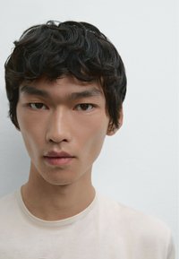 Portrait of a person with short, dark, slightly wavy hair. They wear a light beige crew-neck shirt. Background is a smooth, light color.