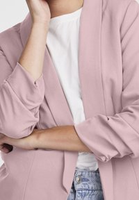 Světle růžový blazer s ohrnutými rukávy a otevřeným designem vpředu, spárovaný s bílou košilí a džíny. Měkká textura a volný střih.