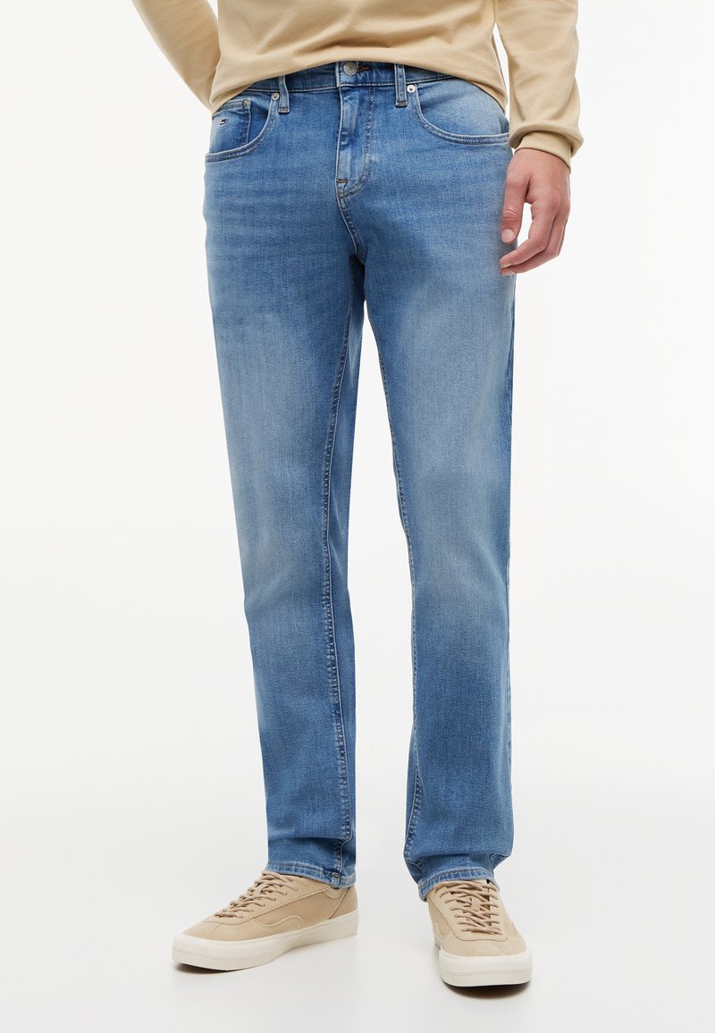 Mann trägt hellblaue Straight-Fit-Jeans, beige Langarmshirt und beige Sneakers und steht vor einem weißen Hintergrund.