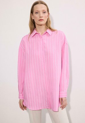 Žena oblečená v voľnej ružovej košeli s dlhými rukávmi a vertikálnymi bielymi pruhmi, stojaca pred jednoduchým svetlým pozadím.