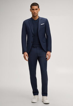 Mann trägt dunkelblauen Anzug mit weißem Einstecktuch, dunkelblauem Hemd und weißen Sneakers, steht vor einfarbigem Hintergrund.