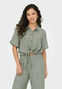 Groen gestreepte shirt met korte mouwen en een geknoopt voorpand, gecombineerd met bijpassende wijde broek. De stof lijkt lichtgewicht en gestructureerd.