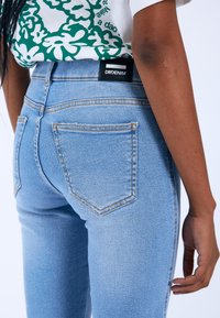 Vaqueros de mezclilla azul claro con una cintura alta, que cuentan con una etiqueta negra, dos bolsillos traseros y costuras naranjas en contraste. Textura suave, forma ajustada.