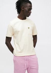 Kremhvid bomulds-T-shirt med korte ærmer, der har en venstre brystlomme og et lille gult broderet logo. Kompletteret med lyserøde joggingbukser.