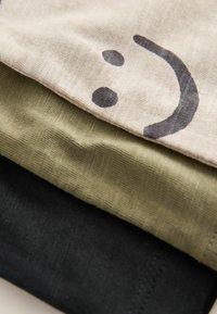 Trois morceaux de tissu empilés : le dessus en beige clair avec un motif de visage souriant, le milieu en vert olive, et le fond en noir, tous avec des surfaces texturées.