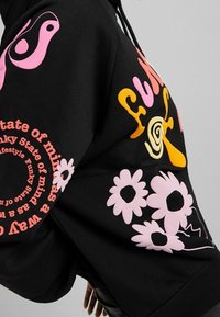 Musta huppari, jossa värikkäitä printtejä: vaaleanpunaisia kukkia, tyyliteltyä tekstiä ja kiemurtelevaa kuviota hihassa. Pehmeä puuvillasekoite ja rento istuvuus.