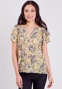 Bloemige blouse met V-hals en korte mouwen, met een geel en zwart bladpatroon op lichte stof. Gepaard met donkere broek.