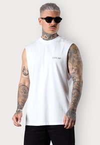 White sleeveless shirt with printed text "ESTEEM" on the chest, paired with black sunglasses. Tattoos visible on the arms. Black pants.