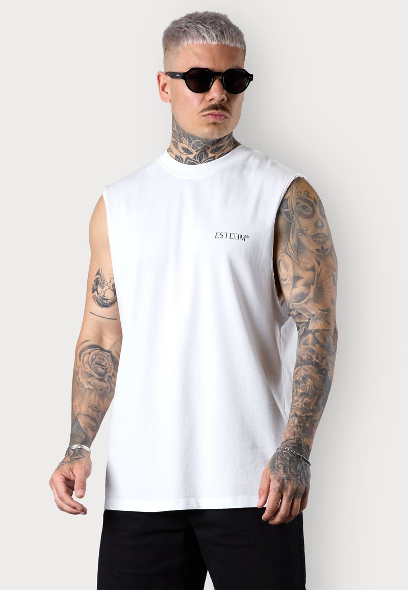 White sleeveless shirt with printed text "ESTEEM" on the chest, paired with black sunglasses. Tattoos visible on the arms. Black pants.